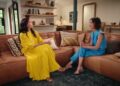 Two women sitting on a brown leather couch, one in a yellow dress and the other in a blue suit.