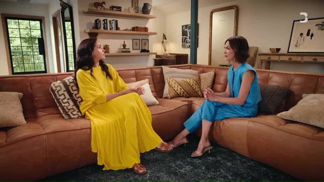 Two women sitting on a brown leather couch, one in a yellow dress and the other in a blue suit.