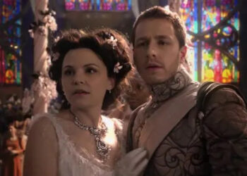 Two people dressed in elaborate, fantasy-style costumes stand together in a setting with stained glass windows, looking concerned