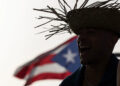 A man wears a straw hat with the Puerto Rican flag in the background before the start of the first show of Puerto Rican singer Bad Bunny’s 30-date concert residency at the Coliseo de Puerto Rico in San Juan, on July 11, 2025. (Credit: Ricardo ARDUENGO / AFP)