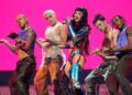 A woman wearing a metallic blue outfit holds a microphone and sings on stage as men dance on either side of her.