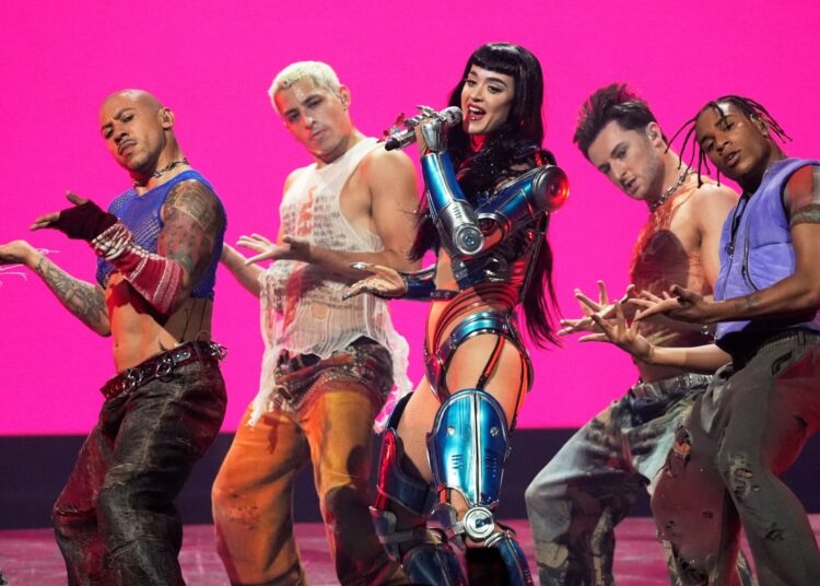 A woman wearing a metallic blue outfit holds a microphone and sings on stage as men dance on either side of her.