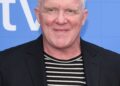 Anthony Michael Hall smiles at a press event, wearing a black blazer over a black-and-white striped shirt.