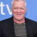Anthony Michael Hall smiles at a press event, wearing a black blazer over a black-and-white striped shirt.