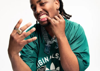 Ash Swaze in a green Adidas shirt, showing off rings and jewellery, with braided hair and a playful expression.