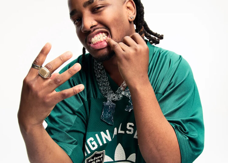 Ash Swaze in a green Adidas shirt, showing off rings and jewellery, with braided hair and a playful expression.