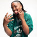 Ash Swaze in a green Adidas shirt, showing off rings and jewellery, with braided hair and a playful expression.