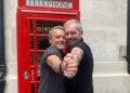 Stefan Dennis and Alan Fletcher dancing in front of a telephone booth.