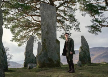 Person in period clothing stands in a circle of stone monoliths, with a mountainous landscape and trees in the background