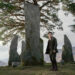 Person in period clothing stands in a circle of stone monoliths, with a mountainous landscape and trees in the background