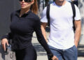 Brooks Nader walks outdoors in Los Angeles wearing black leggings and a fitted zip-up jacket, carrying a gray handbag and holding keys, while Gleb Savchenko follows beside her in a white T-shirt, jeans, and sunglasses.