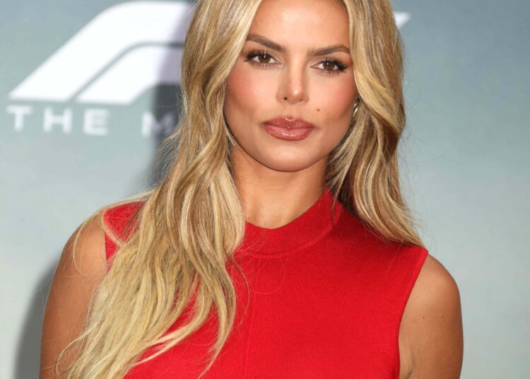Brooks Nader poses at a red carpet event wearing a sleeveless red dress, her long blonde hair styled in loose waves.