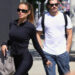Brooks Nader walks outdoors in Los Angeles wearing black leggings and a fitted zip-up jacket, carrying a gray handbag and holding keys, while Gleb Savchenko follows beside her in a white T-shirt, jeans, and sunglasses.