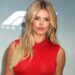 Brooks Nader poses at a red carpet event wearing a sleeveless red dress, her long blonde hair styled in loose waves.