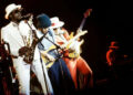A saxophonist and two guitar players stand side-by-side as they perform on stage.