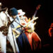 A saxophonist and two guitar players stand side-by-side as they perform on stage.