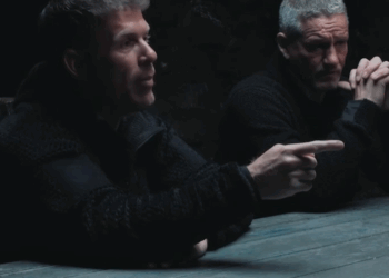 Two men sitting at a table, one pointing.