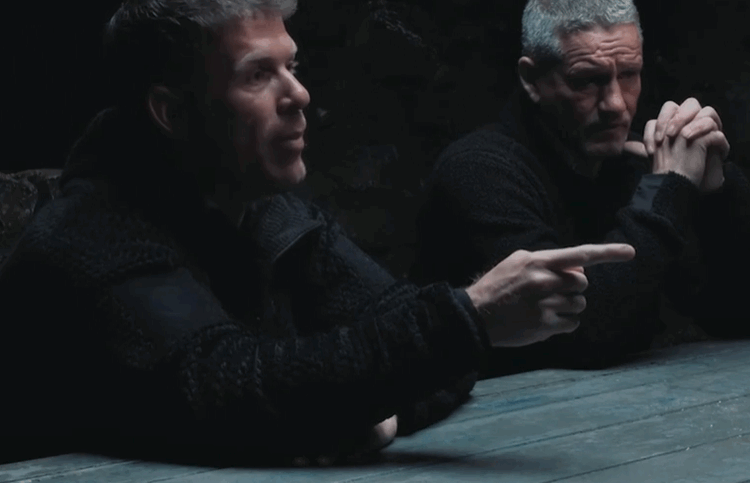 Two men sitting at a table, one pointing.