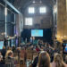 A history talk at an old friary, with a crowd sitting watching two people standing on a stage next to a large screen. The tall stone friary is lit with string lights along the walls.