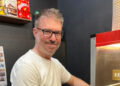 Mr Champion smiles at the camera next to a popcorn making machine. He has short dark hair with some grey in it and he wears a white T-shirt.