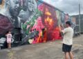 A man takes a photo of his son giving a thumbs up in front of a mural completed in May by artist Steven Teller, depicting the late music producer Johnny Sandlin, surrounded by nine of the many artists he worked with over his career.