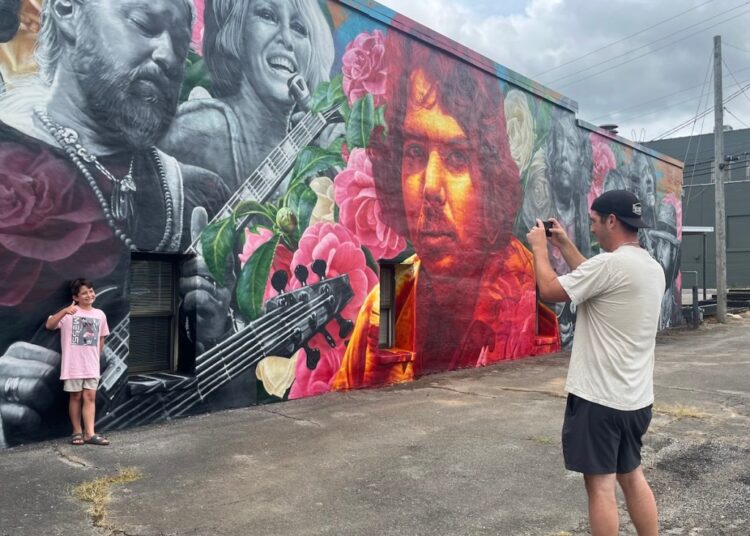 A man takes a photo of his son giving a thumbs up in front of a mural completed in May by artist Steven Teller, depicting the late music producer Johnny Sandlin, surrounded by nine of the many artists he worked with over his career.
