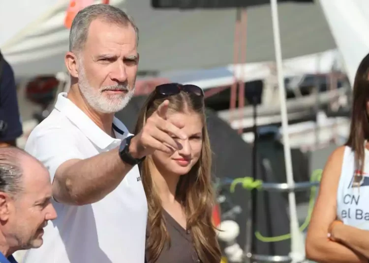 A gray-bearded man points forward while accompanied by several people in an outdoor setting with boats in the background.