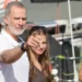 A gray-bearded man points forward while accompanied by several people in an outdoor setting with boats in the background.