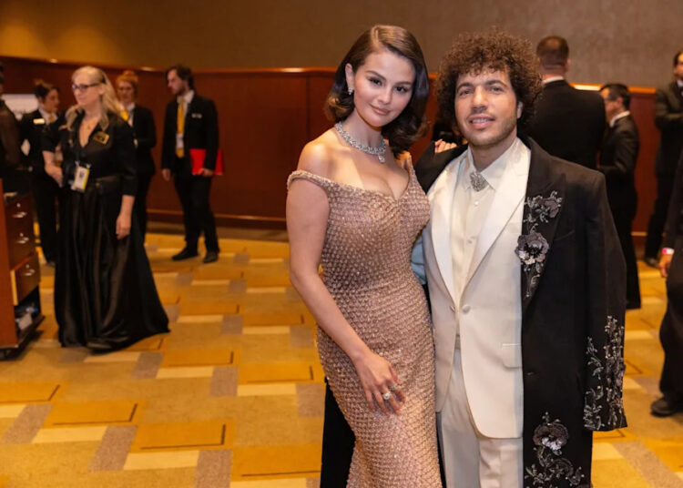 Selena Gomez and Benny Blanco posing together at a formal event, with Gomez in a shimmering champagne-colored gown and Blanco in a white suit with a black floral-embroidered jacket.