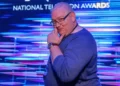 Rick Bronks James Kenny is standing sideways in front of a blue and purple promotional screen at the National Television Awards, with the NTA branding behind him. He is a white man with short grey, thinning hair who is smiling and holding his hand up to his chin in a comedic way. He is wearing a blue jumper, square glasses and a NTA lanyard.