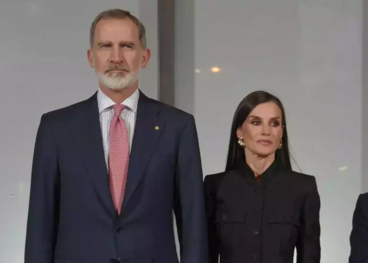 Felipe with a red tie and Letizia dressed in black serious looking to the side
