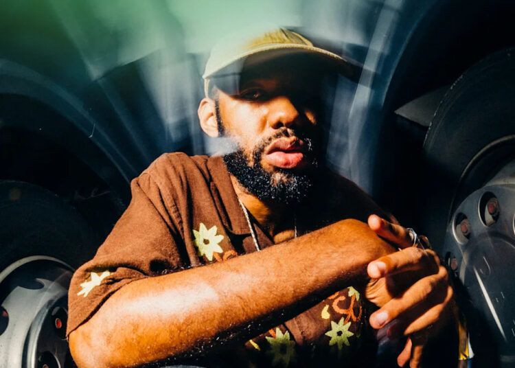 A person with a beard wearing a brown shirt with floral designs and a cap, sitting in front of a blurred background.