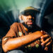 A person with a beard wearing a brown shirt with floral designs and a cap, sitting in front of a blurred background.