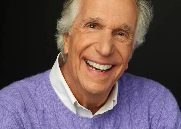 Henry Winkler smiling wearing a purple sweather and a white collared shirt