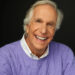 Henry Winkler smiling wearing a purple sweather and a white collared shirt