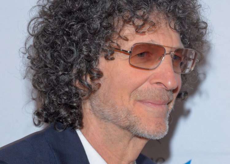Howard Stern in a suit and tie with tinted glasses, smiling slightly at a red carpet event.