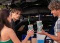 Three people holding smoothies near an open car trunk. Two are facing forward, one is looking back. They appear to be enjoying a casual moment