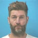 Mugshot of Jay Cutler against a blue background. He has tousled brown hair, a beard, and is wearing a plain light-colored shirt while looking directly at the camera.