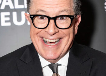 Stephen Colbert smiles at the camera while wearing a suit and tie