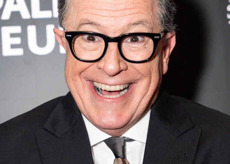 Stephen Colbert smiles at the camera while wearing a suit and tie