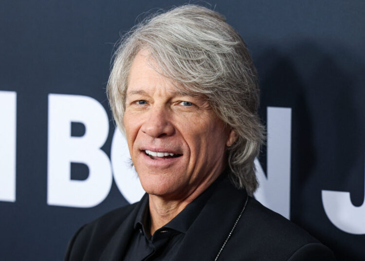 Jon Bon Jovi smiles on the red carpet, wearing a black suit jacket over a black shirt, with his silver hair styled loosely around his face.