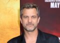Joshua Jackson poses at a red carpet event, wearing a dark blazer with a slightly open collar. He has short gray-streaked hair and light stubble, smiling subtly against a backdrop with yellow and red tones.