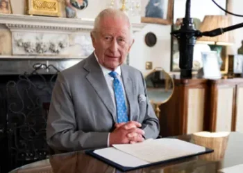 PA Media King Charles III wears a grey suit with a striking blue tie with his hands clasped at the foot of a piece of paper containing the words of his VJ Day message in the Morning Room of Clarence House, London. He is sitting in front of a marble-style fireplace. A microphone hovers near to this face.