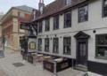 Google A pub with grey walls and dark grey window and door frames is shown with its name sign above the door to the left and an empty seating area in the centre of the frame.