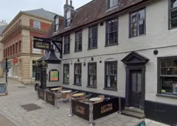 Google A pub with grey walls and dark grey window and door frames is shown with its name sign above the door to the left and an empty seating area in the centre of the frame.