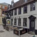 Google A pub with grey walls and dark grey window and door frames is shown with its name sign above the door to the left and an empty seating area in the centre of the frame.