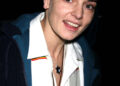 Sinéad O’Connor smiles for the camera, wearing a white shirt with layered necklaces and a dark jacket, her head shaved.