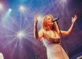 Meg Washington, a woman in her late 30s with long brown hair and wearing a sparkling dress, performs on stage, her arms out.