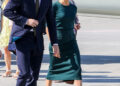 Prince Harry and Meghan Markle walk side by side on a sunny day at an airport. Harry wears a navy suit with a white shirt and tie while Meghan wears a fitted dark green dress with nude heels.