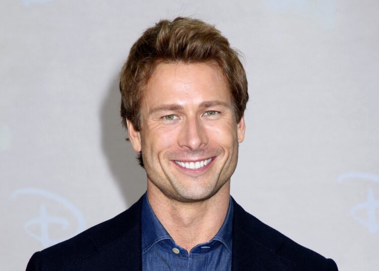 Glen Powell smiles on the red carpet in a navy jacket over a denim button-down shirt